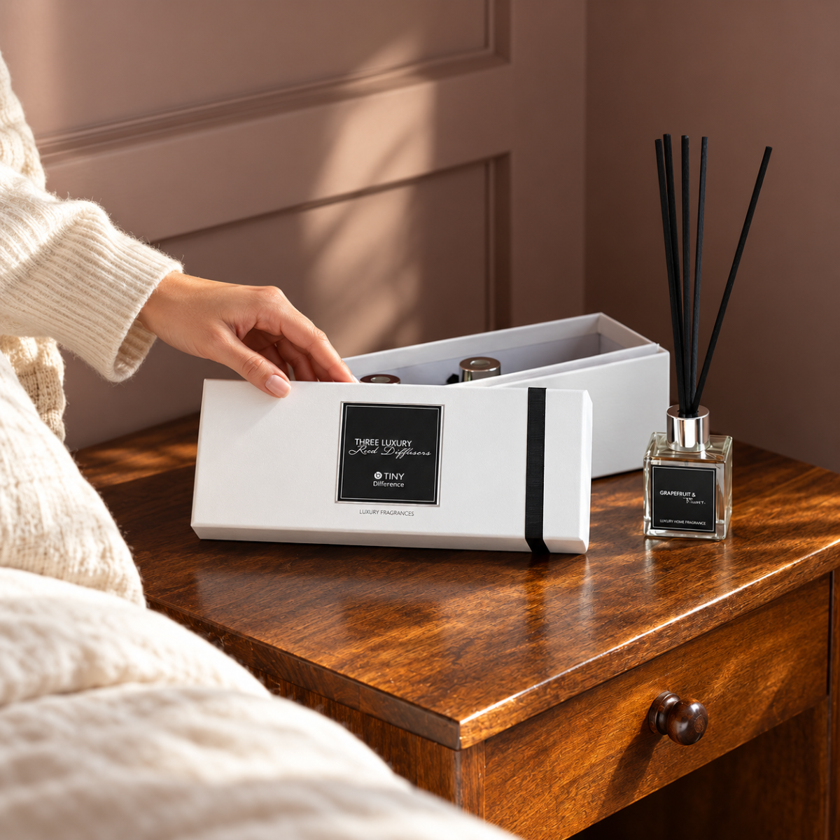 A hand gently opening a white diffuser gift box on a wooden bedside table.